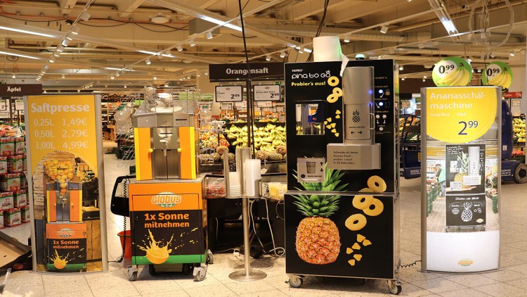 Saftpresse, Ananasschälmaschine und Werbetafel mit Preisen und Motiven von GLOBUS in der Obst- und Gemüseabteilung.