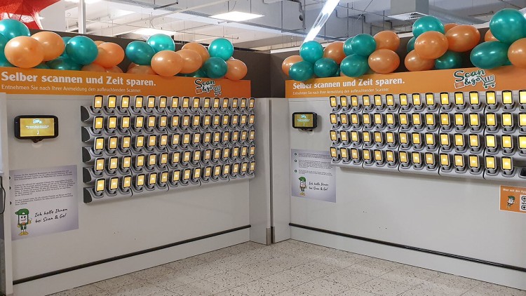 Wand mit zahlreichen Handscannern für „Scan & Go“, orange-grüne Dekoration und Infotafeln mit Anleitung zur Nutzung.