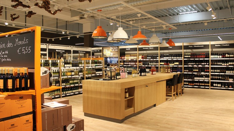 Weinabteilung mit Holzregalen, zentralem Probierstand und einer Tafel mit der Aufschrift „Wein des Monats 5,55 €“.