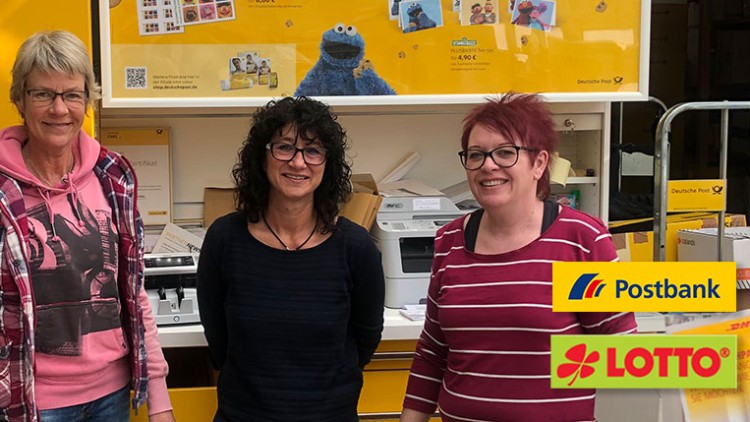 Drei Personen stehen in einer gelben Postfiliale, im Hintergrund Postbank- und Lotto-Logos sowie Pakete und Poster.