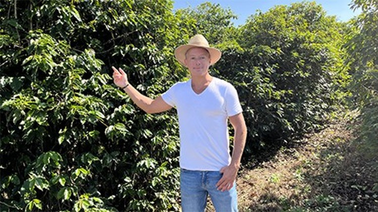 Person in weißem T-Shirt und Strohhut steht vor grünen Pflanzen und zeigt mit einer Hand auf ein Gebüsch.