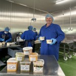 Eine Person in blauer Arbeitskleidung hält Plastikbehälter, im Hintergrund weitere Personen in einer Produktionshalle.