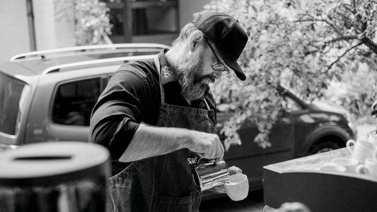 Eine Person mit Schürze und Baseballmütze gießt Flüssigkeit aus einer Kanne in eine Tasse, Autos und Bäume im Hintergrund.