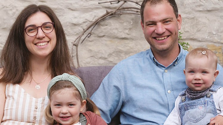 Vier Personen sitzen auf einem Sofa vor einer Steinmauer, darunter zwei Kinder in bunter Kleidung, ein Kind trägt eine Haarspange.