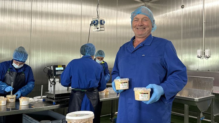 Personen in blauer Schutzkleidung bereiten in einem Industrieraum Lebensmittel zu, eine Person hält zwei verpackte Produkte.