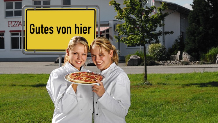 Zwei Personen in weißen Jacken halten eine Pizza vor einem Gebäude mit dem gelben Schildtext „Gutes von hier“.