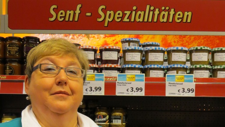 Person in blauer Jacke hält drei Gläser mit Konfitüre vor einem Regal mit der Aufschrift „Senf - Spezialitäten“.