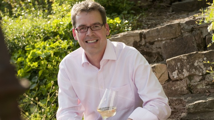 Person in hellem Hemd hält ein Glas mit weißem Wein, sitzt vor einer Naturkulisse mit grünen Büschen und Natursteinstufen.