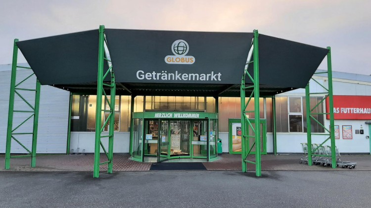 Eingangsbereich eines GLOBUS Getränkemarkts mit grünem Glasdach und Drehkreuz, darüber schwarzes Schild mit Text Getränkemarkt.