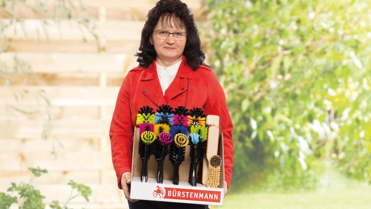Eine Person in roter Jacke hält einen Bürstenträger mit bunten Bürsten und einem Bürstenmann-Logo.