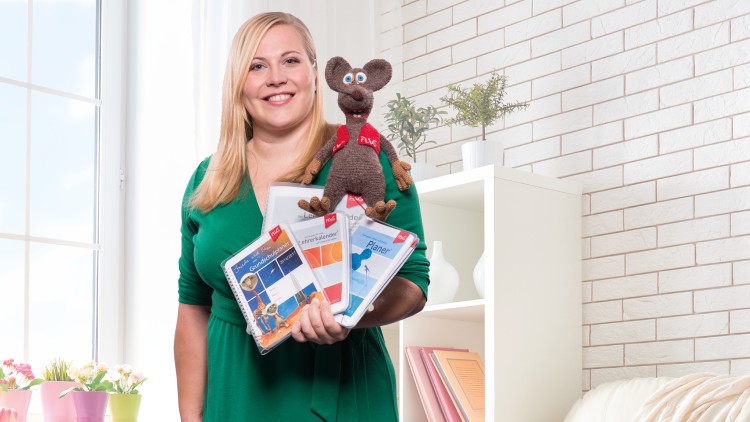 Person in grünem Kleid hält mehrere Planer und Kalender in der Hand, auf denen eine braune Maus mit rotem Halstuch sitzt.