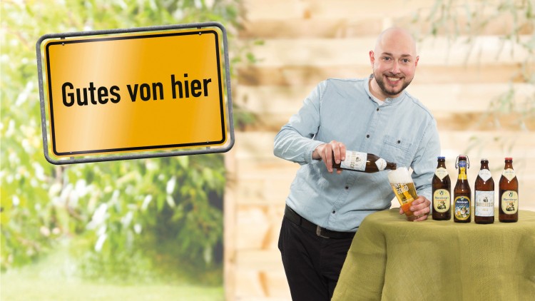 Eine Person schenkt Bier aus einer Flasche in ein Glas ein, daneben stehen fünf Bierflaschen, Schild mit Text „Gutes von hier“.