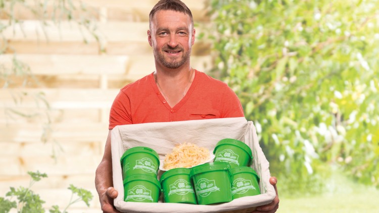 Person in rotem Shirt hält einen Korb mit grün beschrifteten Behältern und geriebenem Sauerkraut, Hintergrund mit Pflanzen und Holz.