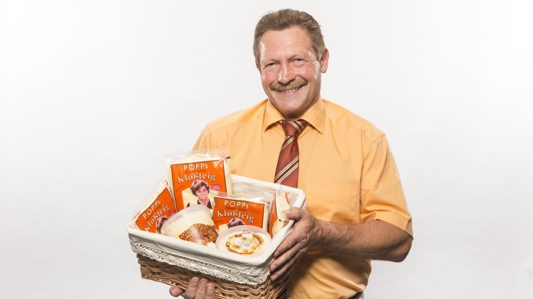 Person in gelbem Hemd hält einen Korb mit mehreren Packungen POPPs Klosteig und anderen Lebensmitteln vor weißem Hintergrund.