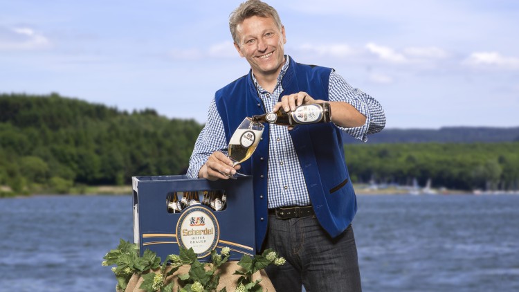 Eine Person im blau karierten Hemd schenkt Scherdel-Bier aus einer Flasche in ein Glas vor einem See mit Wald im Hintergrund.
