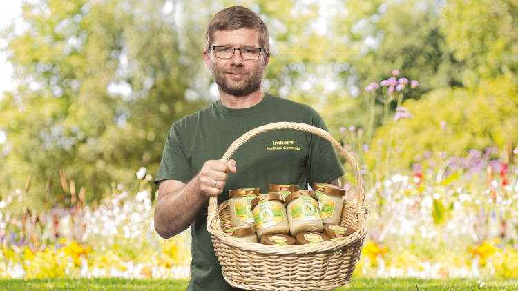 Ein Mensch in grünem Shirt mit der Aufschrift „Imkerei Mathias Gottwald“ hält einen Korb mit mehreren Honiggläsern vor bunter Blumenwiese.
