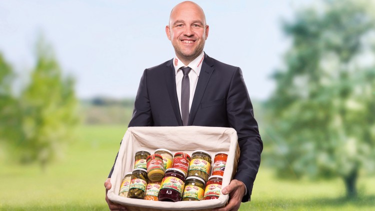 Person in Anzug hält einen rechteckigen Korb mit verschiedenen eingelegten Produkten in Gläsern vor einer grünen Landschaft.