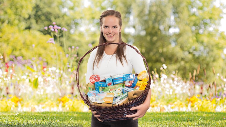 Eine Person hält einen gefüllten Korb mit verschiedenen verpackten Lebensmitteln vor einer blühenden Wiese.