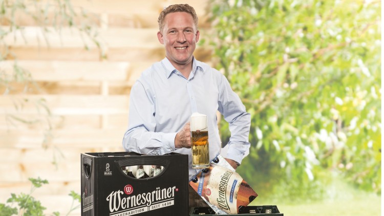 Eine Person in hellblauem Hemd hält ein Bierglas, daneben Bierkisten und eine Bierverpackung der Marke Wernesgrüner.