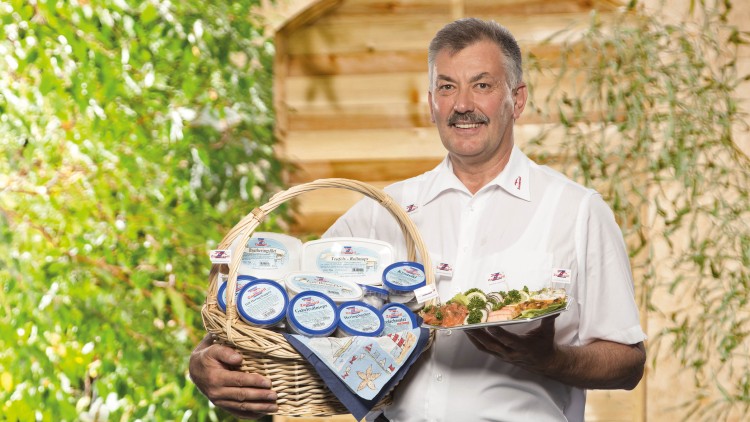 Person in weißem Hemd hält einen gefüllten Präsentkorb mit Fischprodukten und einen Teller mit angerichteten Speisen.
