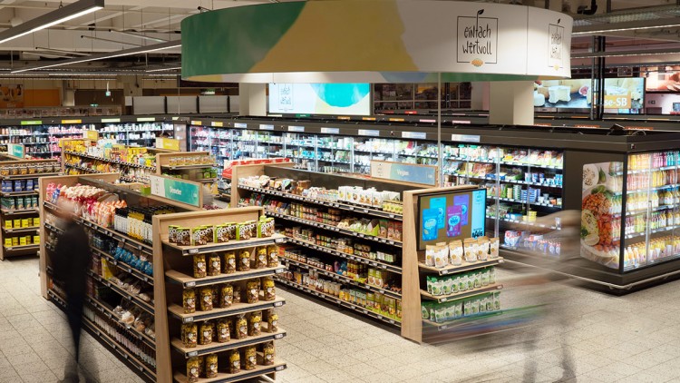 Blick auf einen GLOBUS Supermarkt-Gang mit Regalen für vegane Produkte, einer Kühltheke und einer digitalen Infotafel.