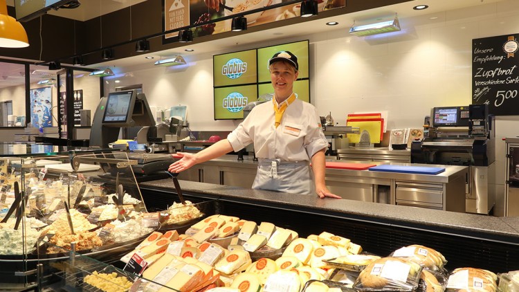 Mitarbeiterin an der GLOBUS-Käsetheke präsentiert eine Auswahl an Käsesorten sowie Feinkost und Brot, im Hintergrund GLOBUS-Logos.