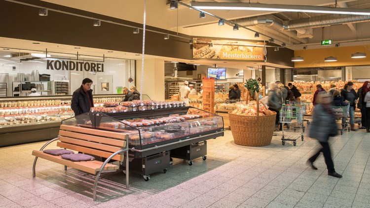 Konditoreiabteilung mit einer Glasvitrine voller Backwaren, einer Holzbank und einem großen Korb mit Broten im Vordergrund.