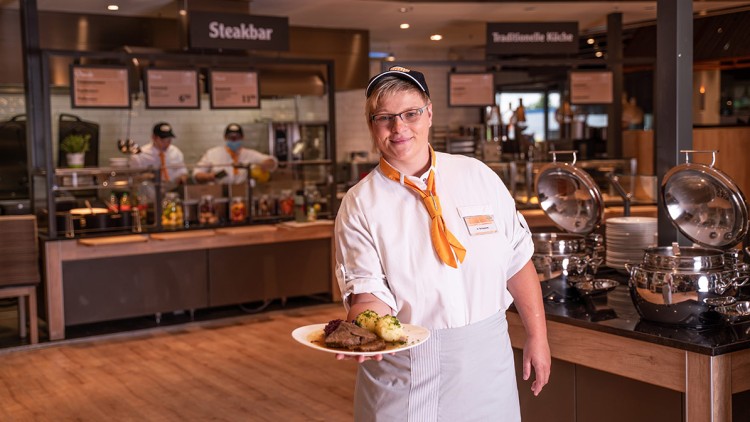 Eine Servicekraft in weißer Uniform mit orangefarbenem Halstuch hält einen Teller mit Fleisch, Soße und Beilagen vor der Steakbar.