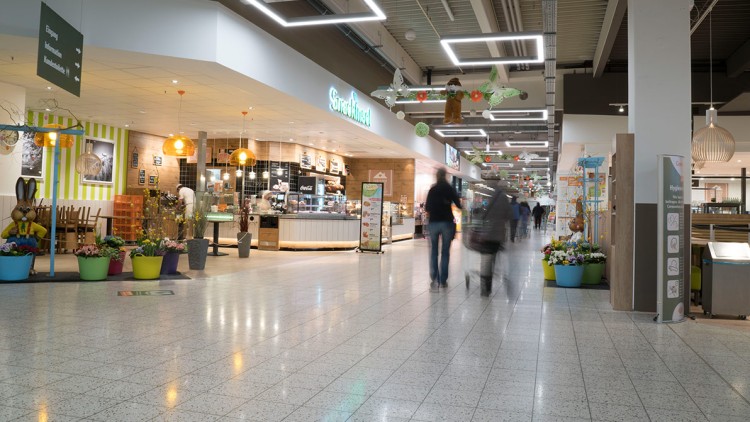 Helle Globus-Marktpassage mit Snackinsel, Sitzbereich, Osterdekoration, Leuchtreklame und vereinzelten Menschen mit Einkaufswagen.