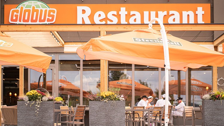 Eingang zum GLOBUS Restaurant mit orangenen Sonnenschirmen, Sitzgelegenheiten und Blumenkübeln auf der Terrasse.