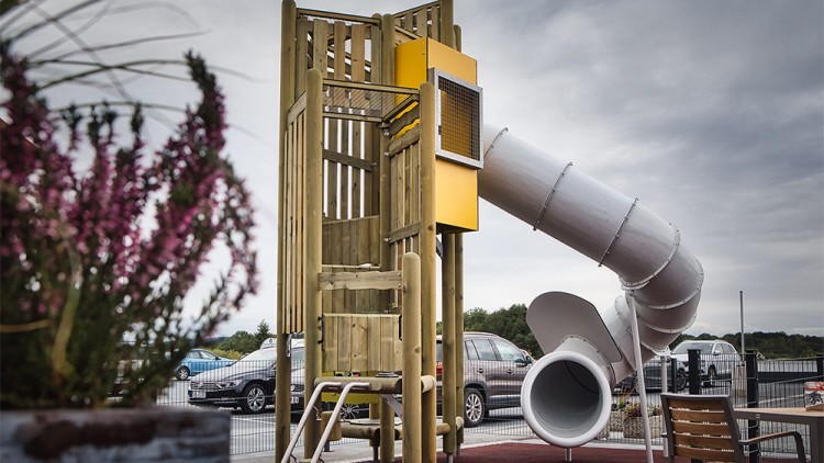Kletterturm mit Rutsche aus Holz und Metall steht auf einer roten Fläche, im Hintergrund geparkte Autos und Pflanzen.