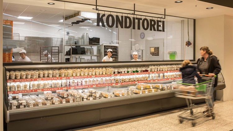 Vor einer Konditorei-Theke mit verpackten Backwaren schiebt eine Person einen Einkaufswagen mit Kind vorbei.