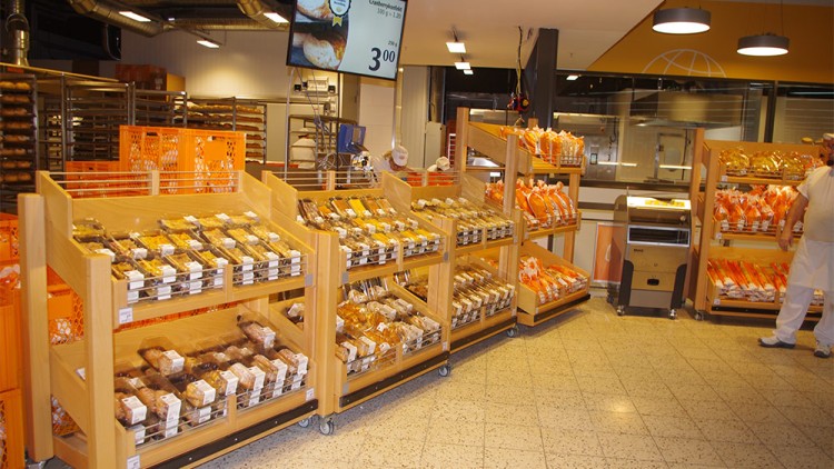 Regale mit abgepacktem Brot und Backwaren stehen in einem Verkaufsbereich, im Hintergrund Backstube und orangene Kisten.