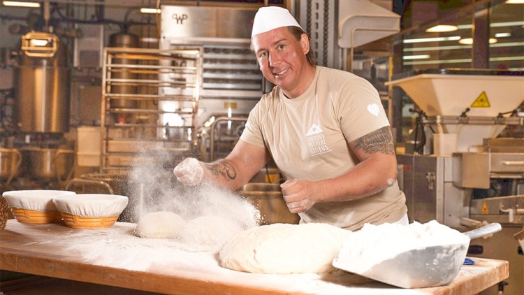 Person bestäubt drei Teiglinge mit Mehl auf einem Holztisch, daneben Körbe und eine große Mehlschaufel, im Hintergrund Bäckerei.