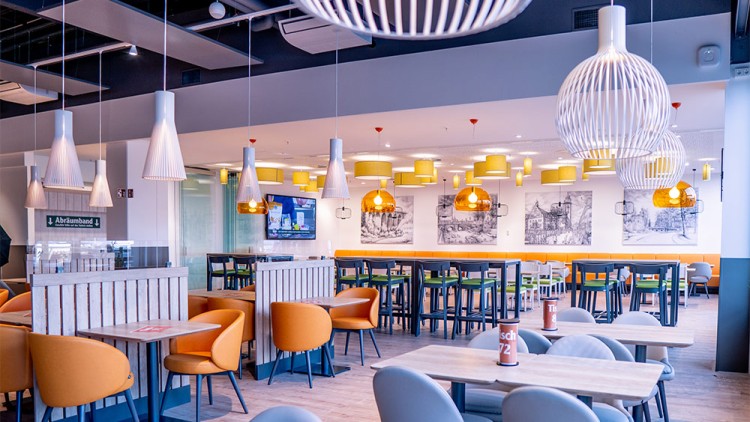Ein modernes Restaurant mit orangefarbenen und grauen Sitzmöbeln, Holztischen, Pendelleuchten und Bildern an der Wand.