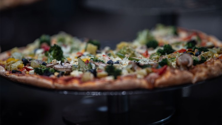 Knusprige Pizza mit bunter Gemüseauswahl wie Brokkoli, Paprika, Oliven, Pilzen und Käse auf einem runden Metallständer.