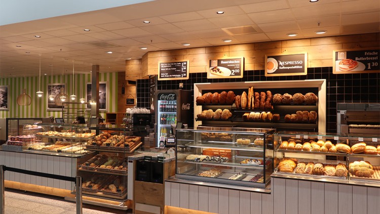 Bäckerei mit verschiedenen Broten, Brötchen und Gebäck in Glasvitrinen, darüber Schilder mit Produktnamen und Preisen.