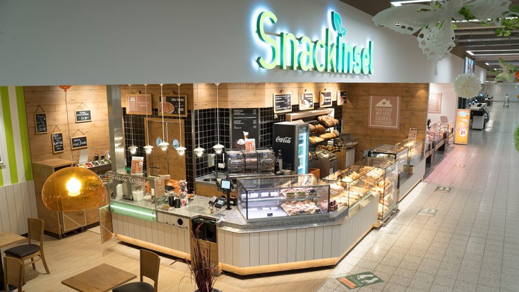 Eine Theke mit Backwaren, Kühlvitrine, Kaffeemaschine und leuchtendem Schriftzug Snackinsel vor Holzwänden und Sitzbereich.