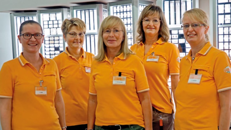 Fünf Personen in orangefarbenen Poloshirts mit GLOBUS-Logo und Namensschildern stehen vor hellen Fenstern.