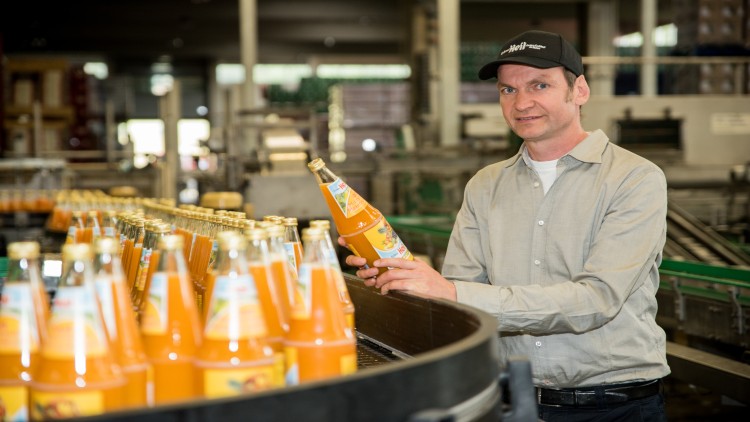 Person in heller Kleidung hält eine Flasche mit oranger Limonade neben einem Förderband voller gleicher Flaschen.