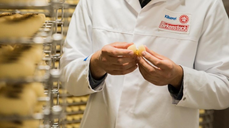 Person in weißem Kittel und Haarnetz hält Käse in den Händen, umgeben von Metallregalen mit gelagerten Käselaiben.