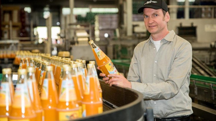 Person in grauem Hemd hält eine gefüllte Flasche Orangensaft vor einer Abfüllanlage mit weiteren Flaschen.