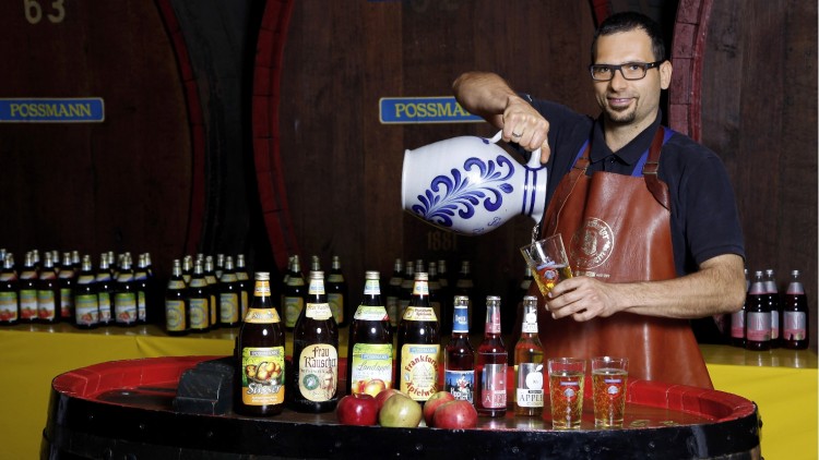 Mann in brauner Schürze schenkt Apfelwein aus einem blau-weißen Krug in ein Glas, Flaschen und Äpfel auf Holzfass davor.