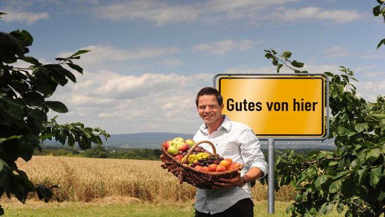 Person hält Korb mit frischem Obst vor gelbem Schild mit schwarzer Aufschrift „Gutes von hier“ in ländlicher Umgebung.