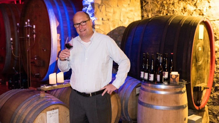 Person in weißem Hemd steht in Weinkeller mit hölzernen Weinfässern, hält ein Glas Rotwein, Kerzen und Flaschen im Hintergrund.