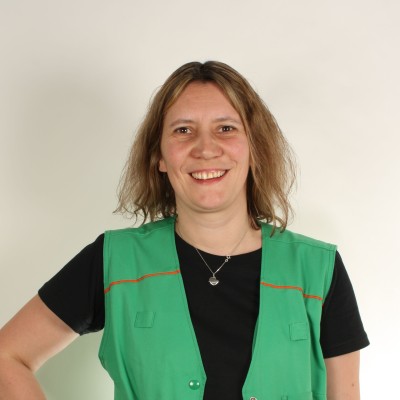 Person in schwarzem T-Shirt trägt grüne Weste mit orangefarbenen Akzenten und mehreren Taschen vor hellem Hintergrund.