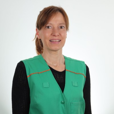 Person trägt eine grüne Weste mit orangen Nähten und schwarzem Oberteil vor einem neutralen Hintergrund.