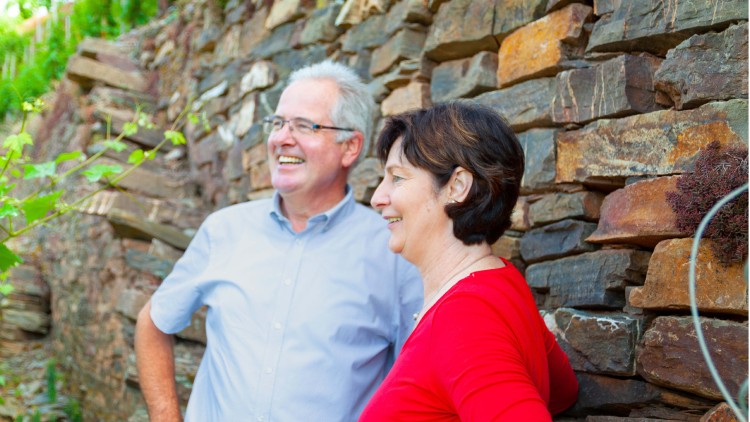 Zwei Personen stehen vor einer Mauer aus Natursteinen, im Hintergrund sind grüne Pflanzen zu sehen.