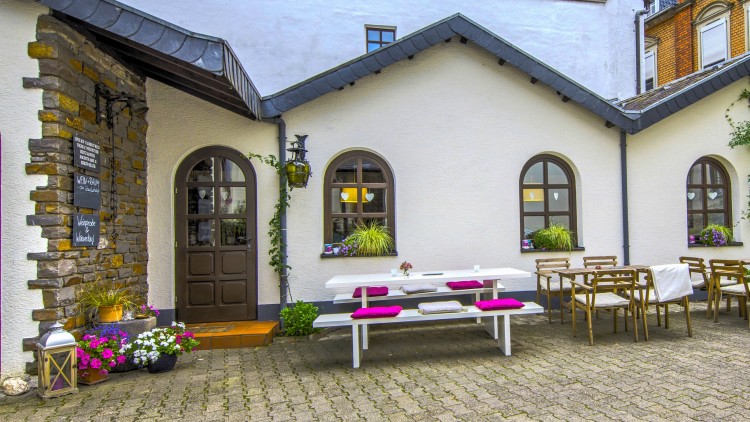 Innenhof mit weißer Hausfassade, Holztüren, Fenstern, Holztischen, Stühlen, weißer Sitzbank, Pflanzen und einer Tafel mit Tagesinfo.