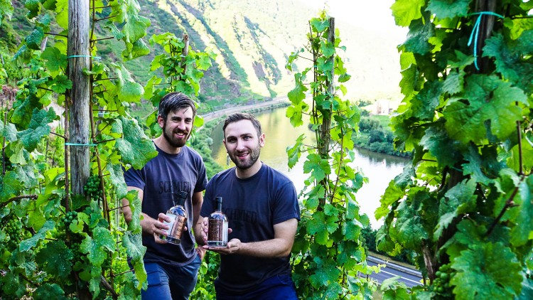 Zwei Personen halten Getränkeflaschen in einem Weinberg mit Blick auf einen Fluss und grüne Hügellandschaft.